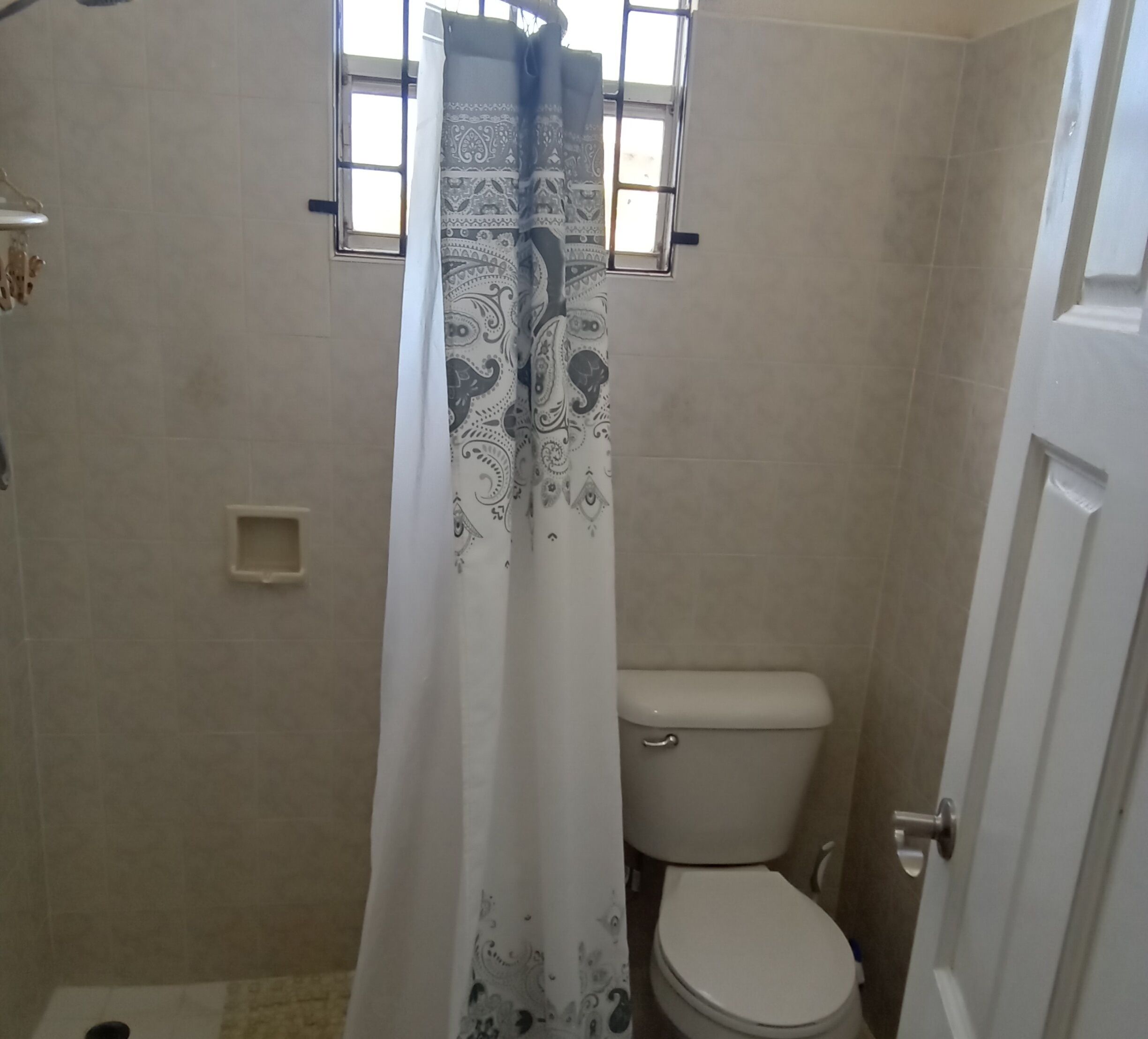 A bathroom with a toilet, a shower curtain with a pattern, and a window with bars.
