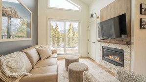 A modern living room with a sofa, ottomans, a fireplace, a TV, and a balcony with a mountain view. - Summit View Penthouse in Downtown Canmore at Brookside Lane (Canmore)