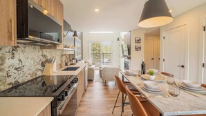 A modern kitchen with a marble countertop, wooden cabinets, and a dining area with a table set for four. - Summit View Penthouse in Downtown Canmore at Brookside Lane (Canmore)