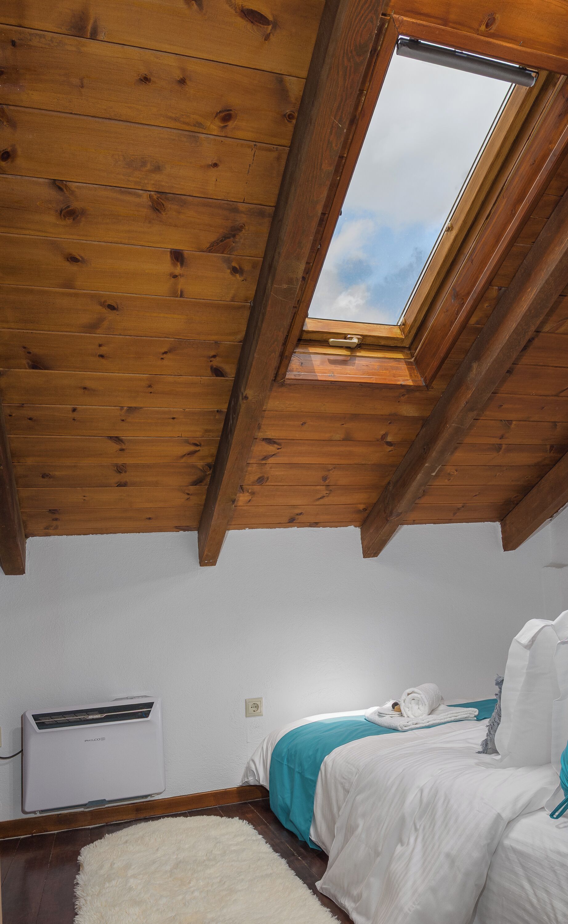 A room with a sloped wooden ceiling, a skylight, a bed with white bedding and a teal accent, and a white radiator.
