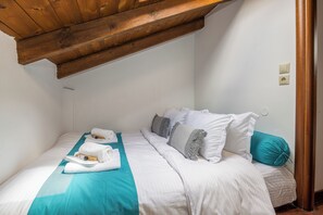 Une chambre avec un plafond en bois, des murs blancs, un lit avec une literie blanche et des touches de couleur turquoise, et une fenêtre donnant sur un paysage.