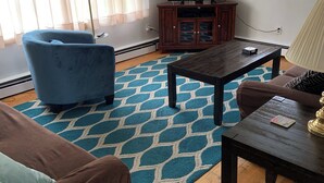 A living room with a blue armchair, a brown sofa, a coffee table, a television on a wooden cabinet, and a patterned rug. - Cozy 3-bedroom cottage in Old Orchard Beach (Old Orchard Beach)