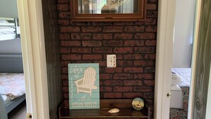 A room with a brick wall, a framed picture, a sign with a chair, a small shelf, and a bed in the background. - Cozy 3-bedroom cottage in Old Orchard Beach (Old Orchard Beach)