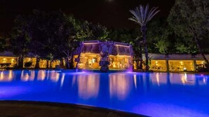 A nighttime view of a hotel pool with a waterfall feature, surrounded by trees and illuminated by ambient lighting.
