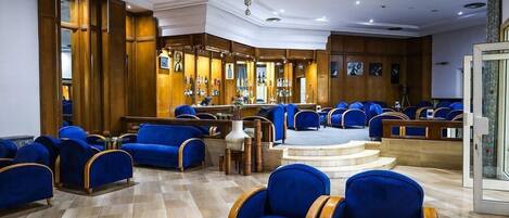 A hotel lobby with blue upholstered seating, a bar area, and a staircase leading to an upper level.