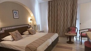A hotel room with a bed, a small table, a chair, a lamp, and a curtain.