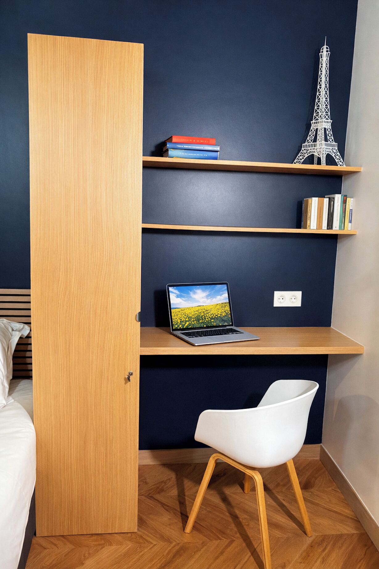 Une chambre avec une porte en bois, un lit, un bureau avec un ordinateur portable, des étagères remplies de livres et une maquette de la Tour Eiffel, ainsi qu’une chaise blanche.