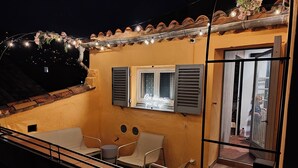 Un balcón con mesa y sillas, una ventana con contraventanas y luces colgantes.