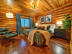 Un dormitorio de estilo cabaña de madera con una cama, una silla, una ventana con cortinas, un candelabro y cuadros enmarcados en la pared.
