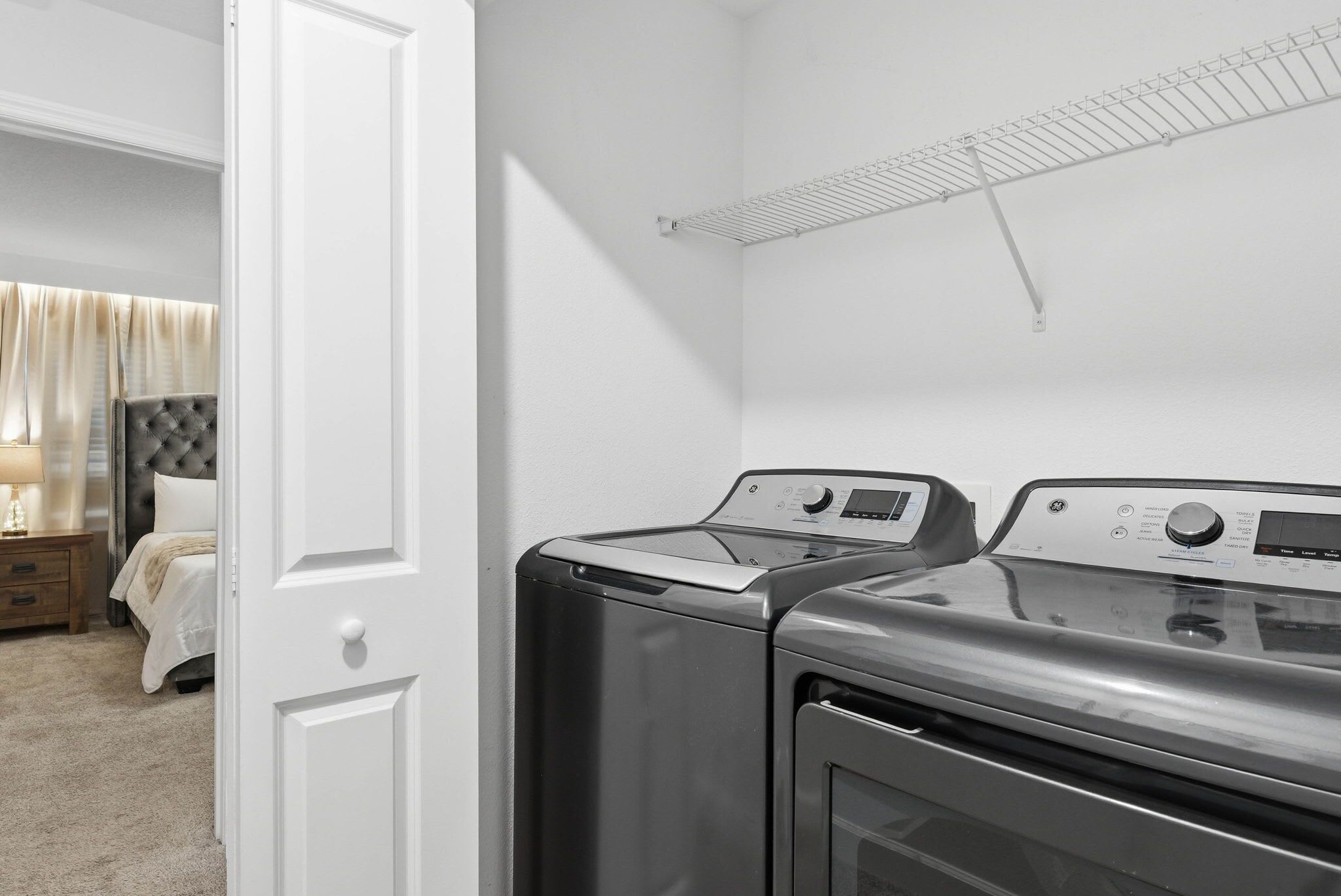 A laundry room with two front-loading washers and dryers, a white door, and a bed with a headboard in the adjacent room.
