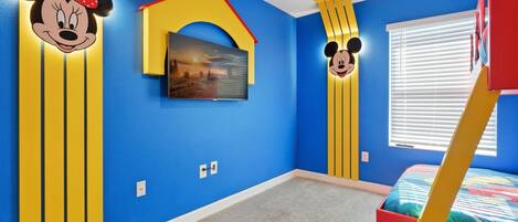 A room with blue walls, yellow vertical stripes, a Mickey Mouse cutout, a bed with a colorful bedspread, and a window with blinds.