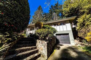 A two-story house with a stone staircase leading to the front door, surrounded by lush greenery and trees. - Cozy Forest Retreat in Horseshoe Bay | Walk to Beaches & Village (West Vancouver)