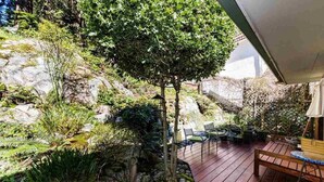 Una terraza de madera con mobiliario exterior, un árbol pequeño y un área de jardín rocoso.