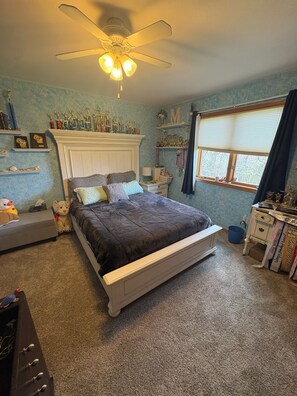 Une chambre à coucher avec un lit, un ventilateur de plafond, une fenêtre avec des rideaux et des étagères où sont exposés des trophées.