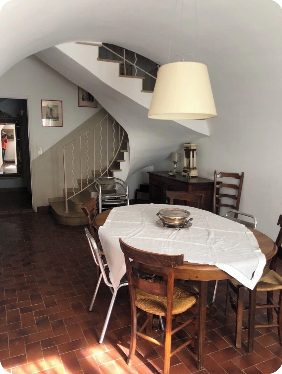A dining area with a round table, chairs, a lamp, a staircase, and a wall with framed pictures.