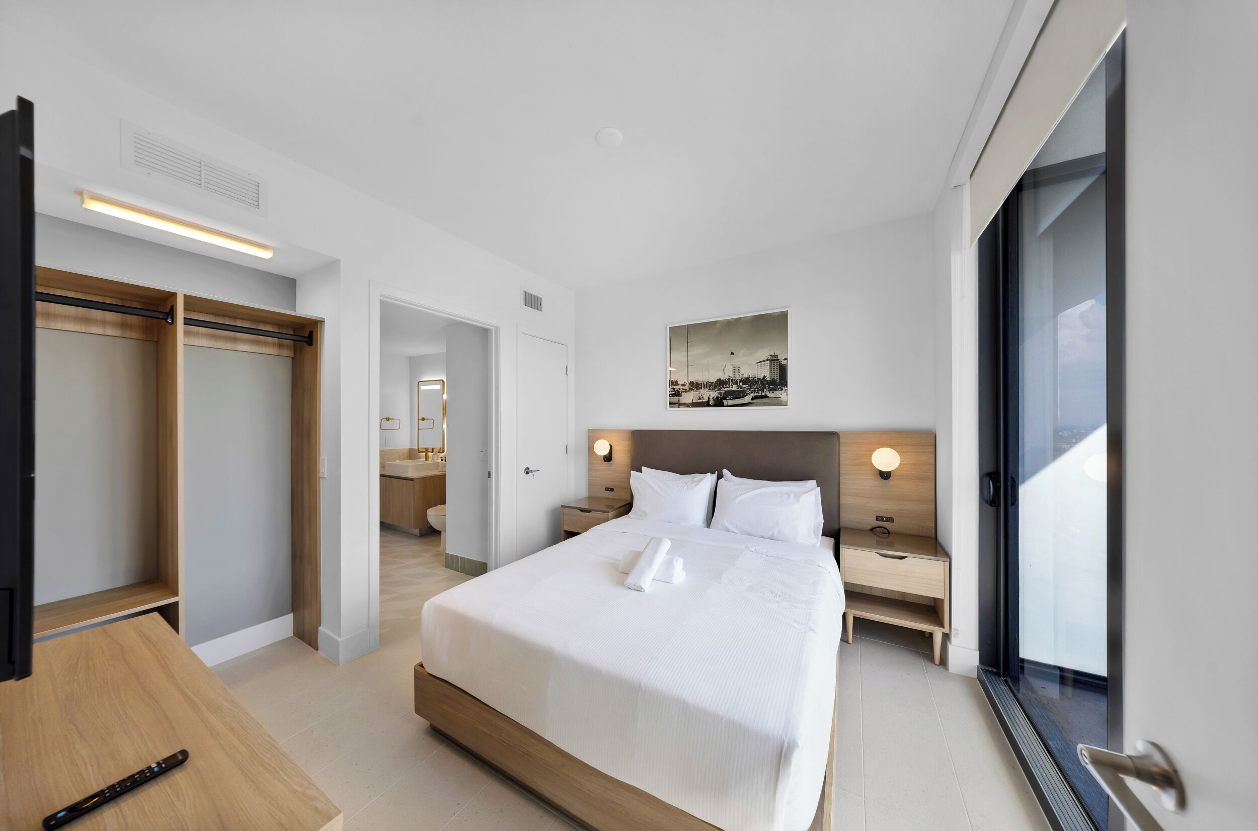 A hotel room with a bed, a wooden desk, a wardrobe, a nightstand, a wall-mounted light, and a window with a cityscape view.