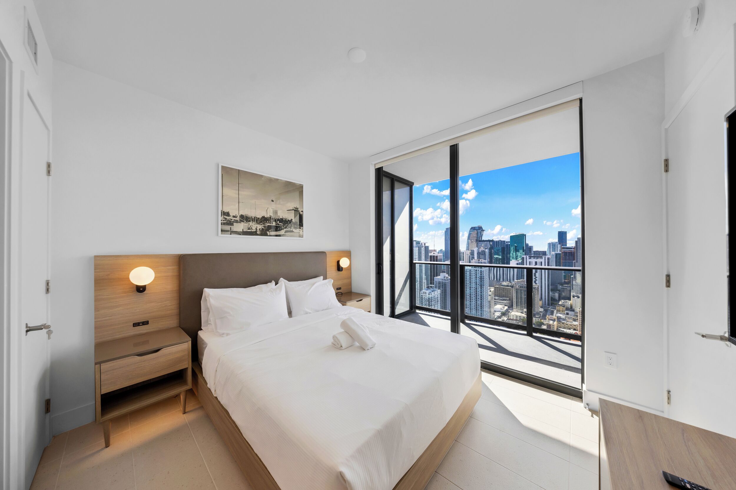 Une chambre d’hôtel moderne dotée d’un grand lit, d’une table de chevet, d’une lampe fixée au mur et offrant une vue sur la ville grâce à de grandes fenêtres.