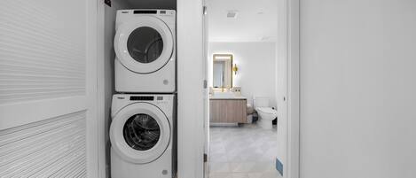 Un cuarto de lavado con dos lavadoras apiladas, un baño con inodoro y lavamanos, y un pasillo que conduce a un dormitorio.