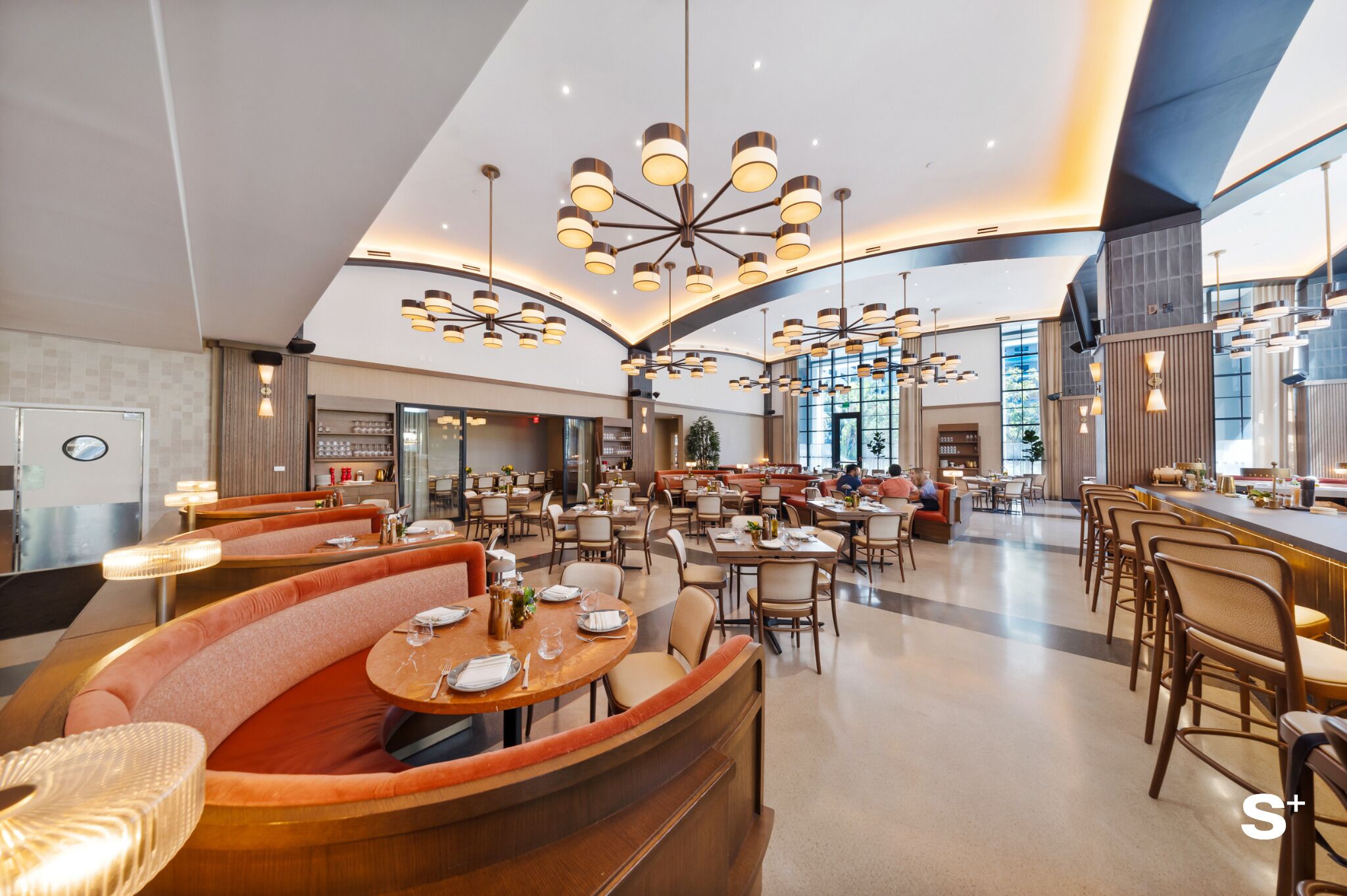 Un hall d’hôtel spacieux, doté de nombreux espaces de détente, notamment des banquettes et des tables, ainsi que d’un grand lustre.