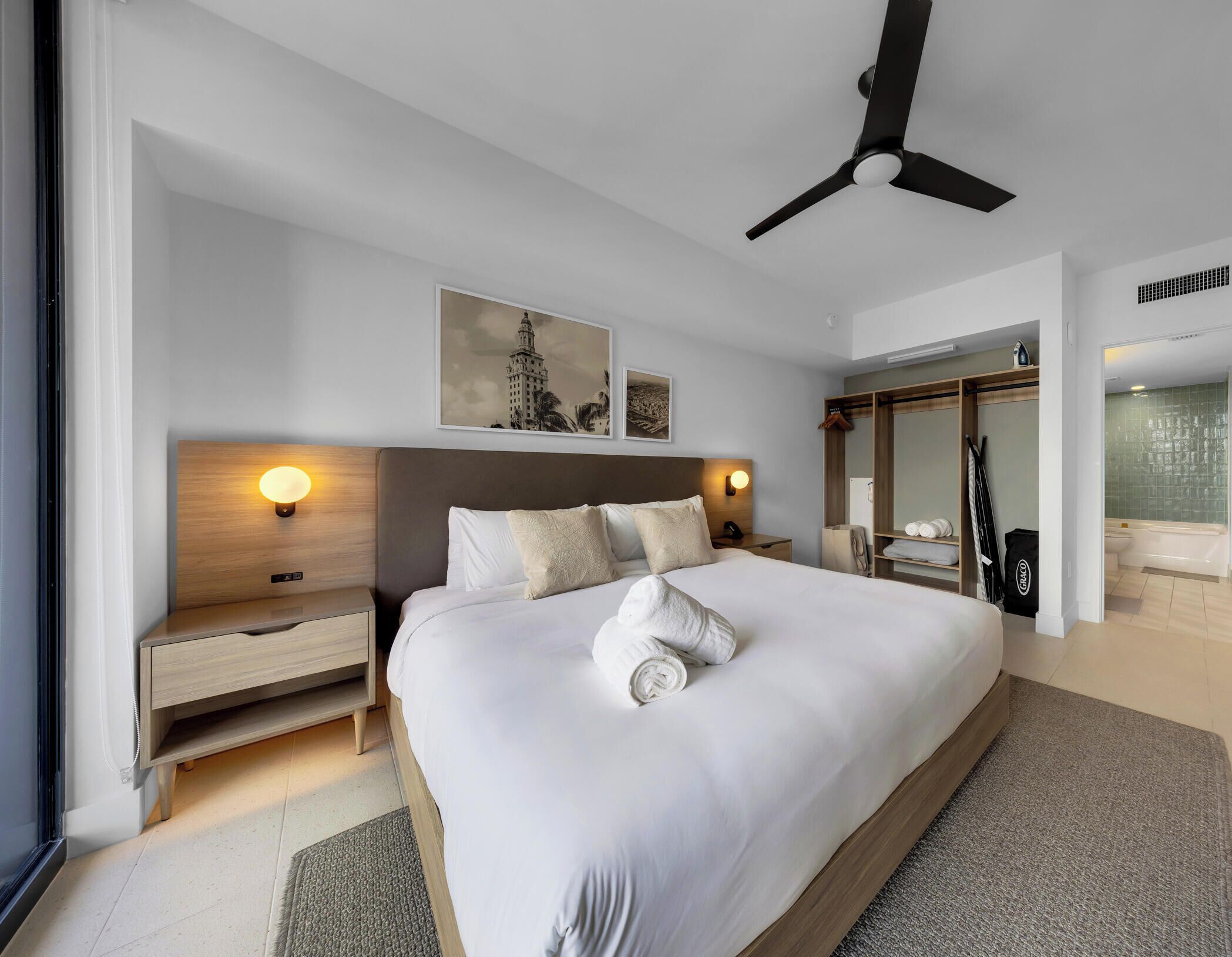 A modern hotel room with a large bed, bedside tables, a ceiling fan, artwork on the wall, and a bathroom visible through an open door.