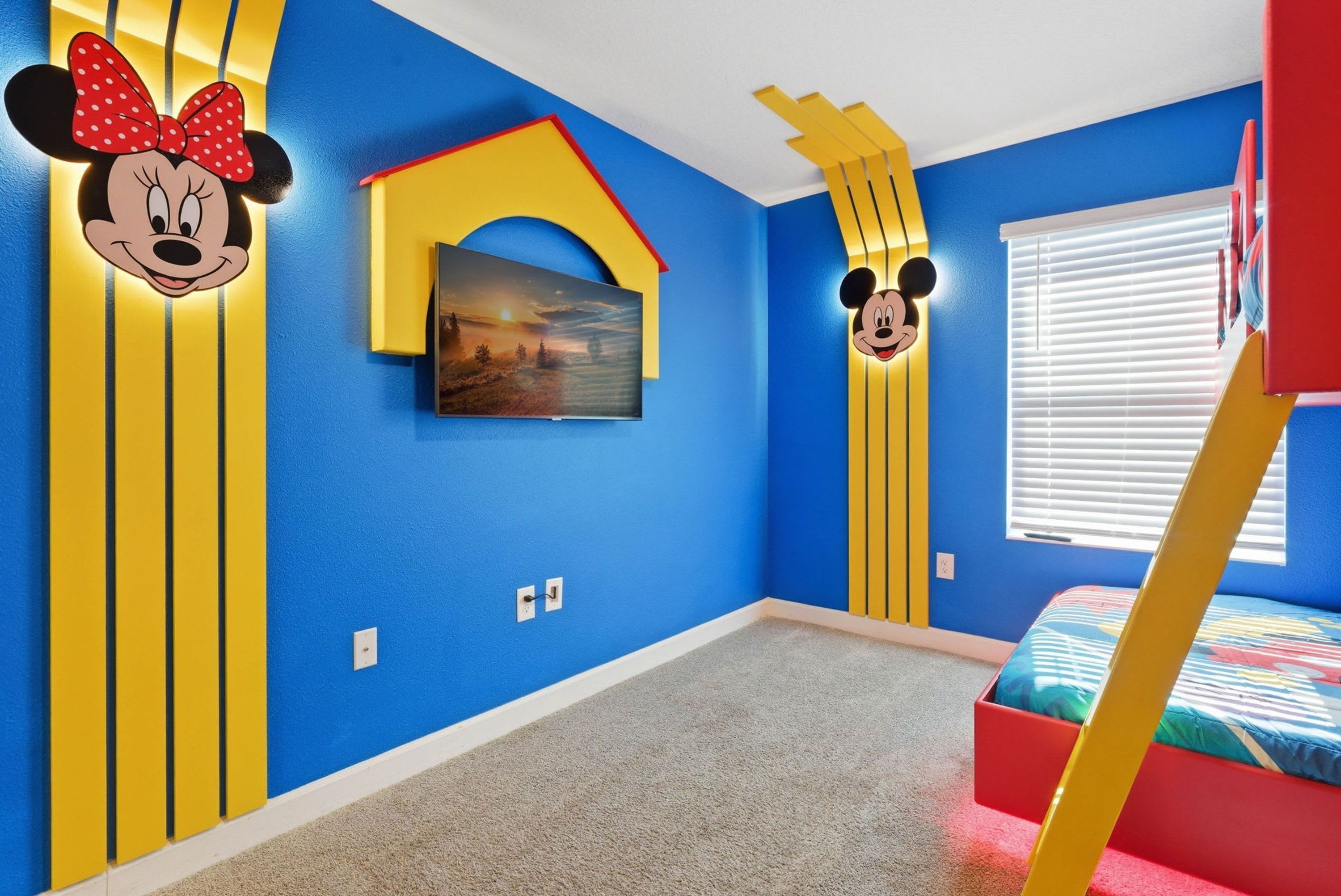 Une pièce aux murs bleus, décorée de rayures verticales jaunes, ornée d’une tête de Mickey Mouse, avec un lit recouvert d’une courtepointe colorée et une fenêtre munie de stores.