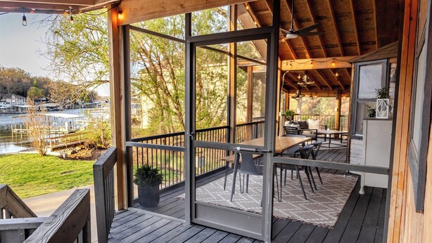Una terraza de madera con cerramiento de vidrio, mobiliario exterior y vista a un lago.