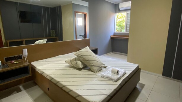 A hotel room with a bed, a bedside table, a mirror, a TV, and a window with a view of greenery. - Hotel Paraty (São Paulo)