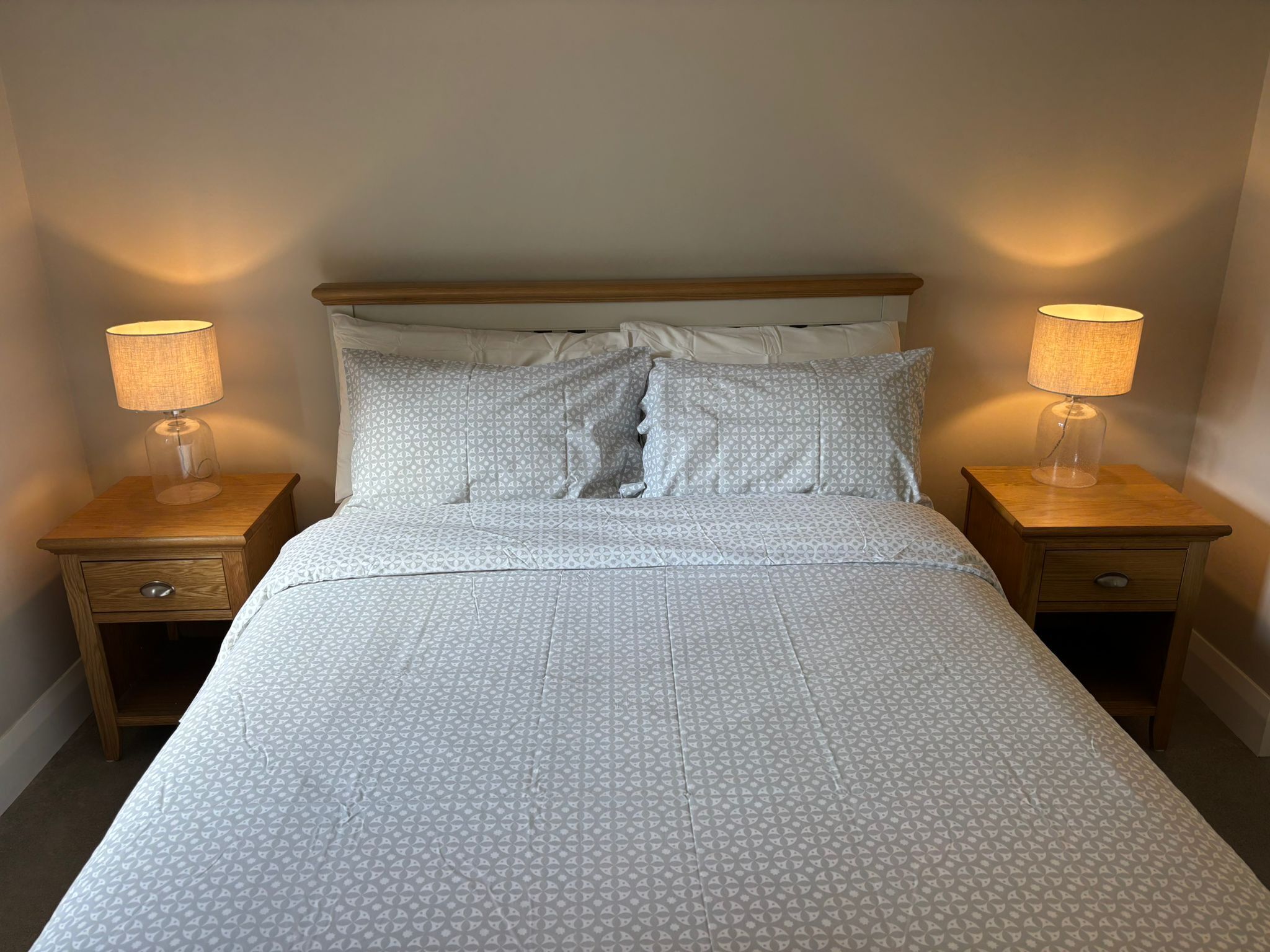 Cama bem arrumada com colcha estampada, duas luminárias de cabeceira e cabeceira de madeira.