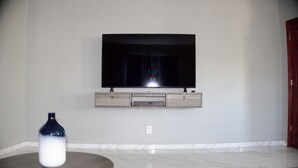 Uma sala de estar com televisão de tela plana na parede, uma mesa redonda de madeira com um vaso e um tapete estampado.