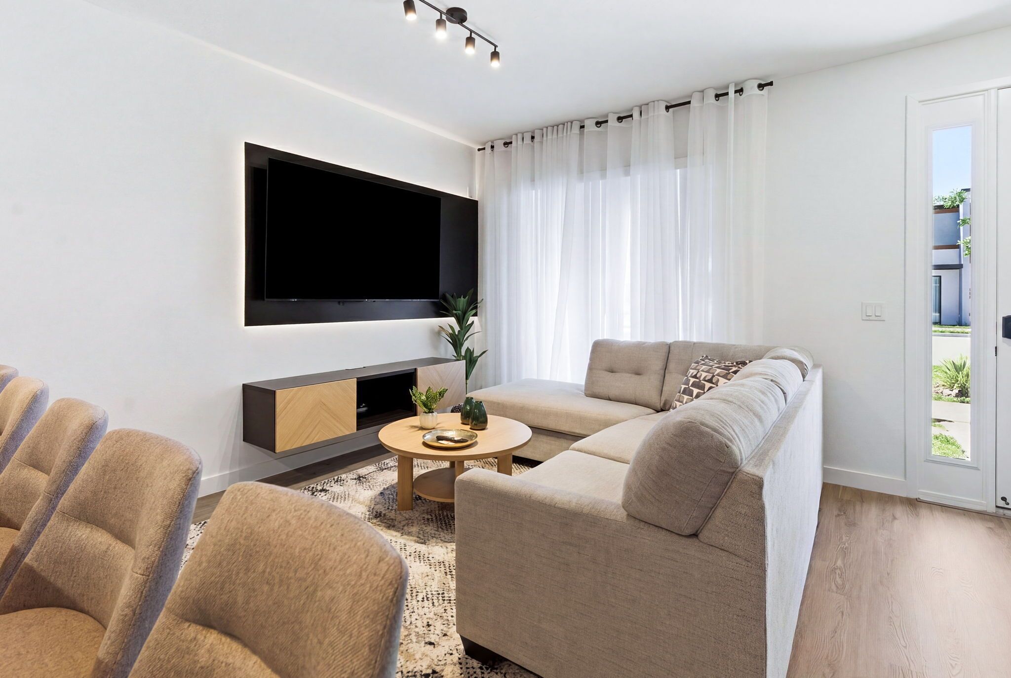 Ein modernes Wohnzimmer mit einer Couch, Sesseln, einem Couchtisch, einem an der Wand montierten Flachbildfernseher und einem großen Fenster mit Vorhängen.