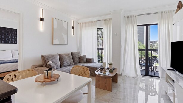 A modern living room with a sofa, a dining table, a TV, and a balcony with curtains. - NEW Cozy Apt | Near Universal, Icon Park & IDrive (6165-1304) (Orlando)