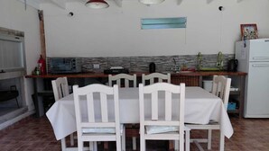 Un coin repas avec une table et des chaises blanches, une cuisine en arrière-plan et un sol carrelé.