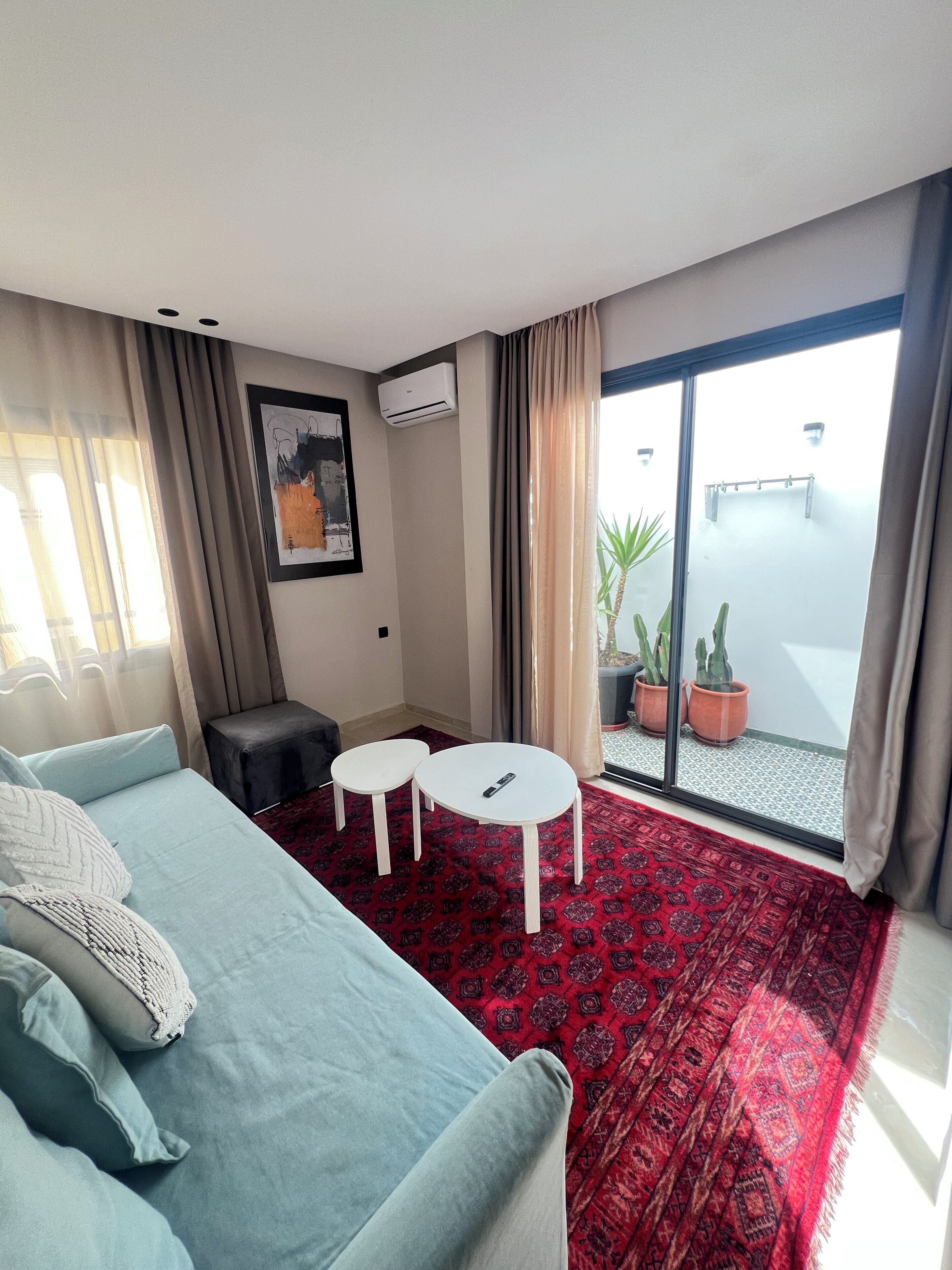 A hotel room with a bed, two small tables, a sofa, a red patterned carpet, a large window with curtains, and a potted plant.