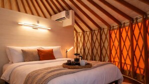 A cozy bedroom with a bed, pillows, a bedside table with a coffee pot, and a wall-mounted air conditioner. - Goldfinch Yurt - Scenic Mountain Retreat (Rising Fawn)