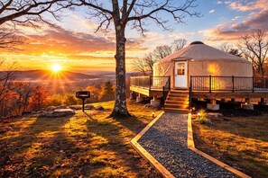 Eine Jurte mit einer Holzterrasse und einem Kiesweg, der davor führt, umgeben von Bäumen und mit Blick auf einen Sonnenuntergang.