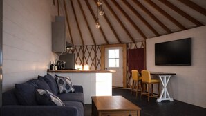 A cozy living room with a sofa, coffee table, and a TV. There's a kitchen area with a window and a dining table with chairs. - Goldfinch Yurt - Scenic Mountain Retreat (Rising Fawn)