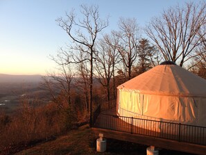 Eine Jurte mit Holzterrasse und Metallgeländer, umgeben von Bäumen und Blick auf die Landschaft.