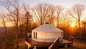Eine Jurte mit einer Holzterrasse und Geländer, umgeben von Bäumen und einem Sonnenuntergang im Hintergrund.