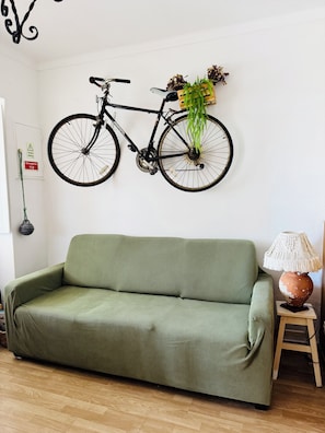 Ein grünes Sofa, ein am Wand befestigtes Fahrrad, ein kleiner Tisch mit einer Lampe und ein an der Wand angebrachtes Notzeichen.