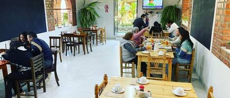 A dining area with wooden tables and chairs, a TV screen, and potted plants.