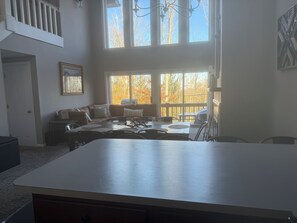 Amplia sala de estar con una gran mesa de comedor, un conjunto de sofás y vistas a follaje otoñal a través de amplias ventanas.