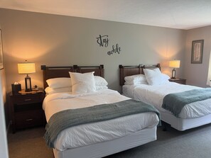 A hotel room with two beds, each with a wooden headboard, white bedding, and a green throw blanket. There are bedside lamps on each nightstand.