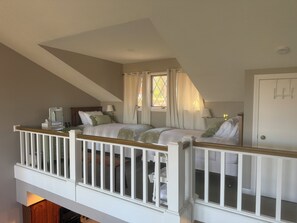 Un dormitorio con techo inclinado, una cama con ropa blanca, una mesita de noche, un espejo, una ventana con cortinas y una puerta con manija.