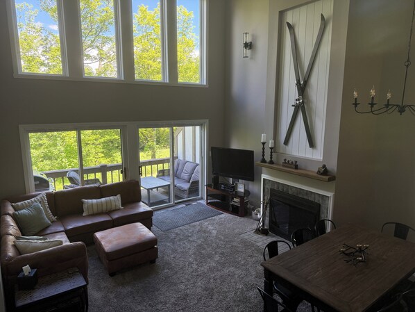 A spacious living room with a fireplace, a television, and a large window overlooking a deck.