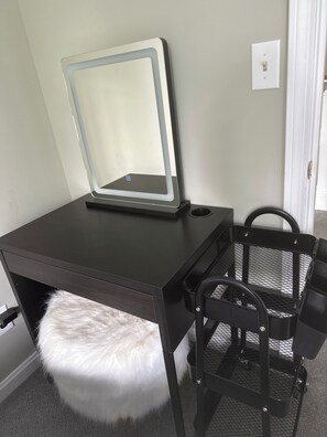 Un escritorio de madera oscura con un espejo, un taburete blanco y mullido, y una silla negra con respaldo de malla.