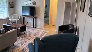Een woonkamer met een televisie, een witte stoel, een donkerne fauteuil, een kamerbreed tapijt en een plafondventilator.