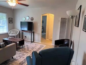 A living room with a television, a white chair, a dark armchair, a patterned rug, and a ceiling fan.