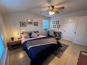 A bedroom with a bed, bedside table, lamp, ceiling fan, desk, chair, and framed pictures on the wall.