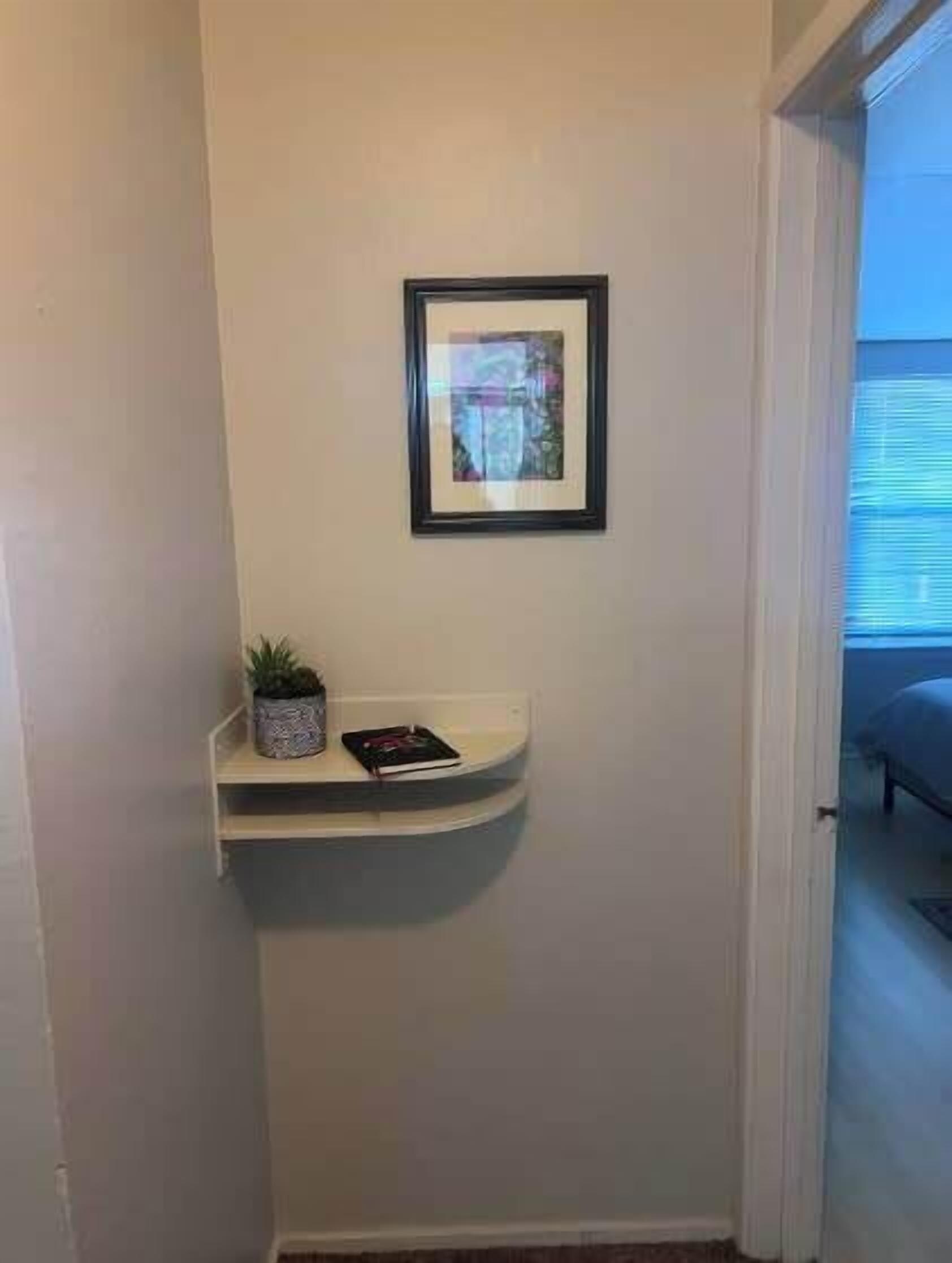 A hallway with a white wall, a framed picture, a small shelf with a plant and a book, and a bed in the adjacent room.