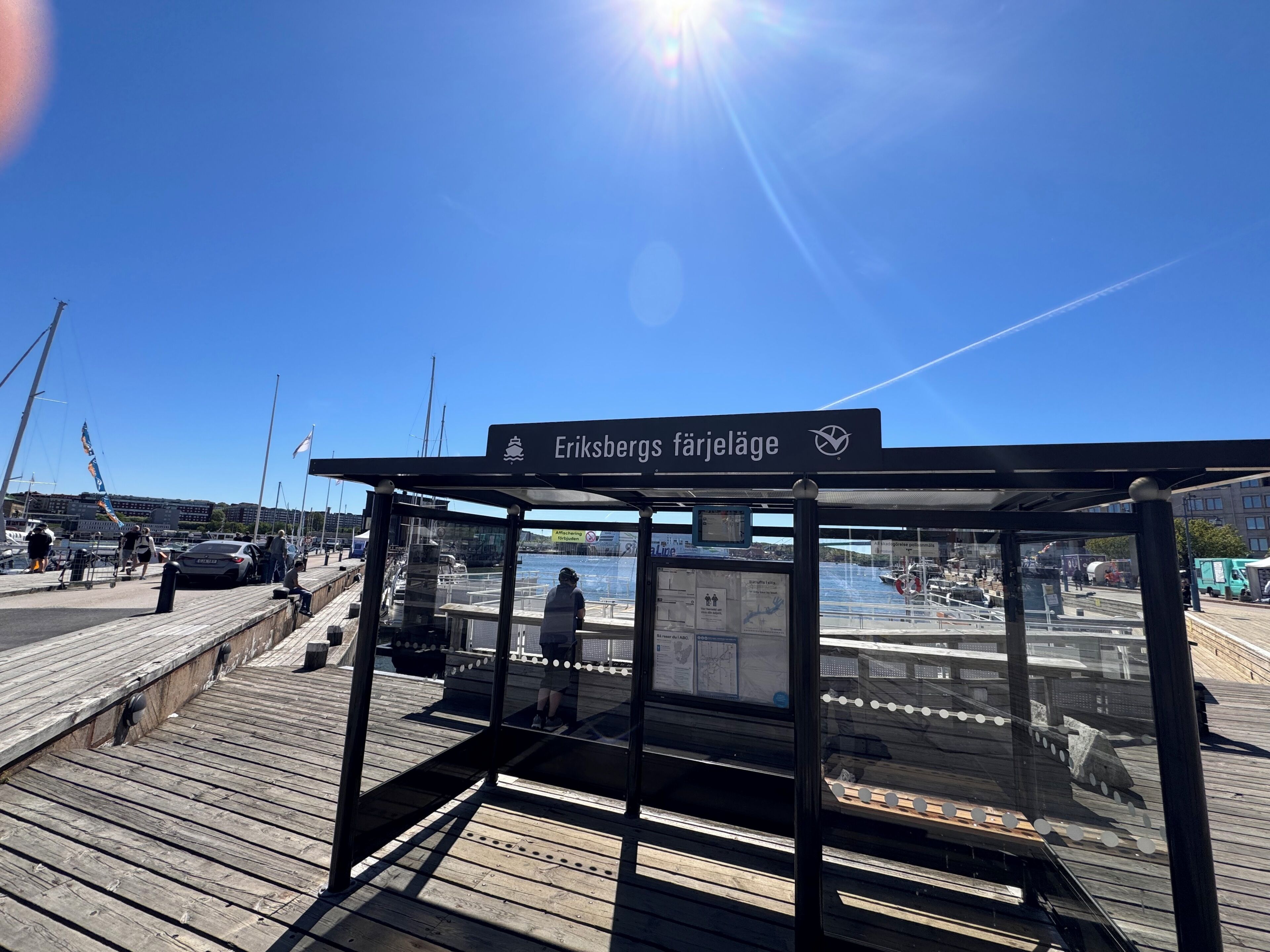 Eine Holzsteg mit einer geschützten Sitzgelegenheit, einem Schild mit der Aufschrift 'Eriksbergs färjeläge' und Blick auf Boote und Wasser.